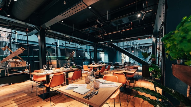 A modern restaurant with wooden tables and orange chairs, featuring a staircase and large windows. - Clarion Hotel Mestari (Helsinki)