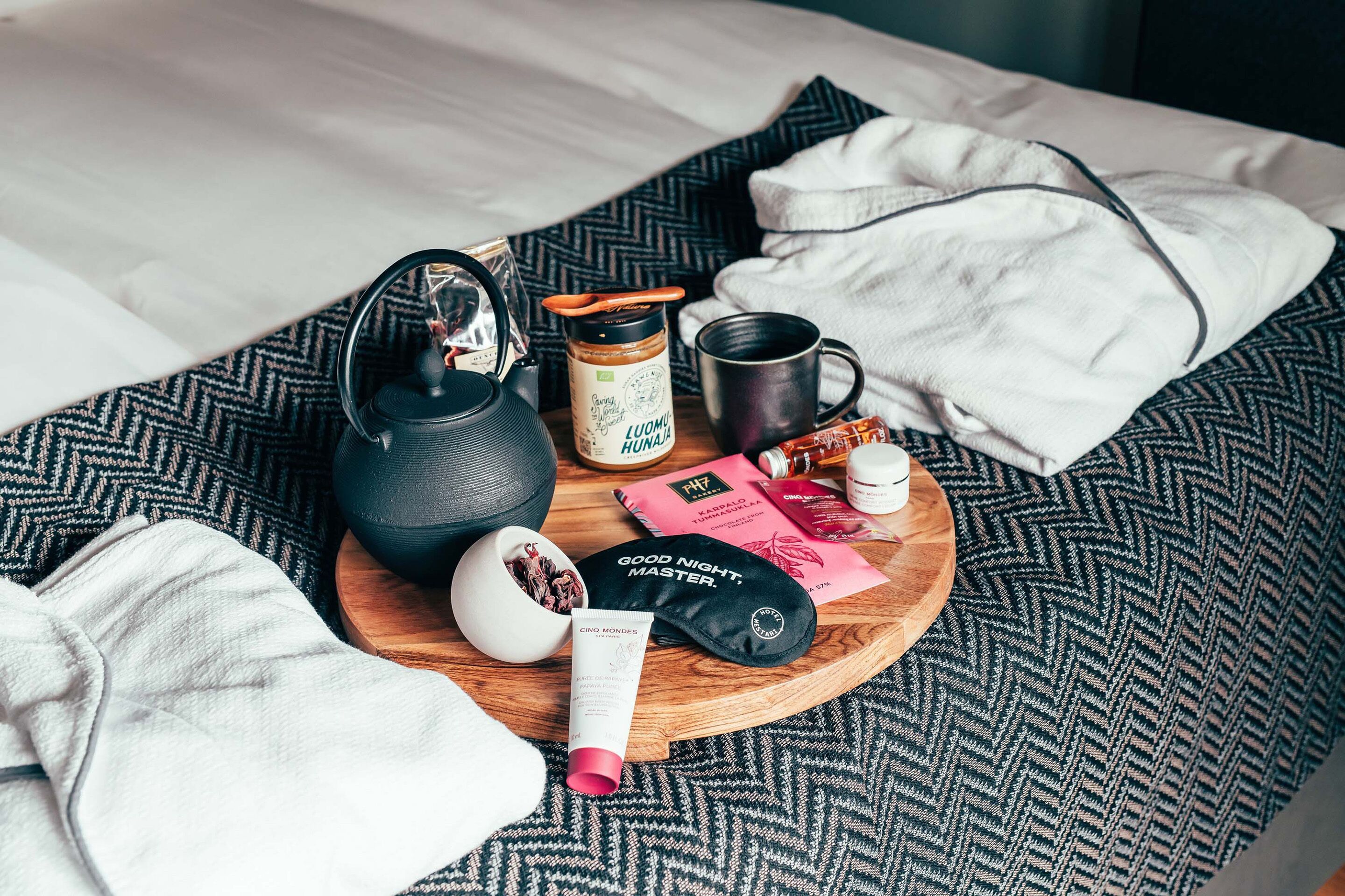 A neatly arranged breakfast tray on a bed with a patterned bedspread, including a kettle, mug, jar, and various breakfast items.