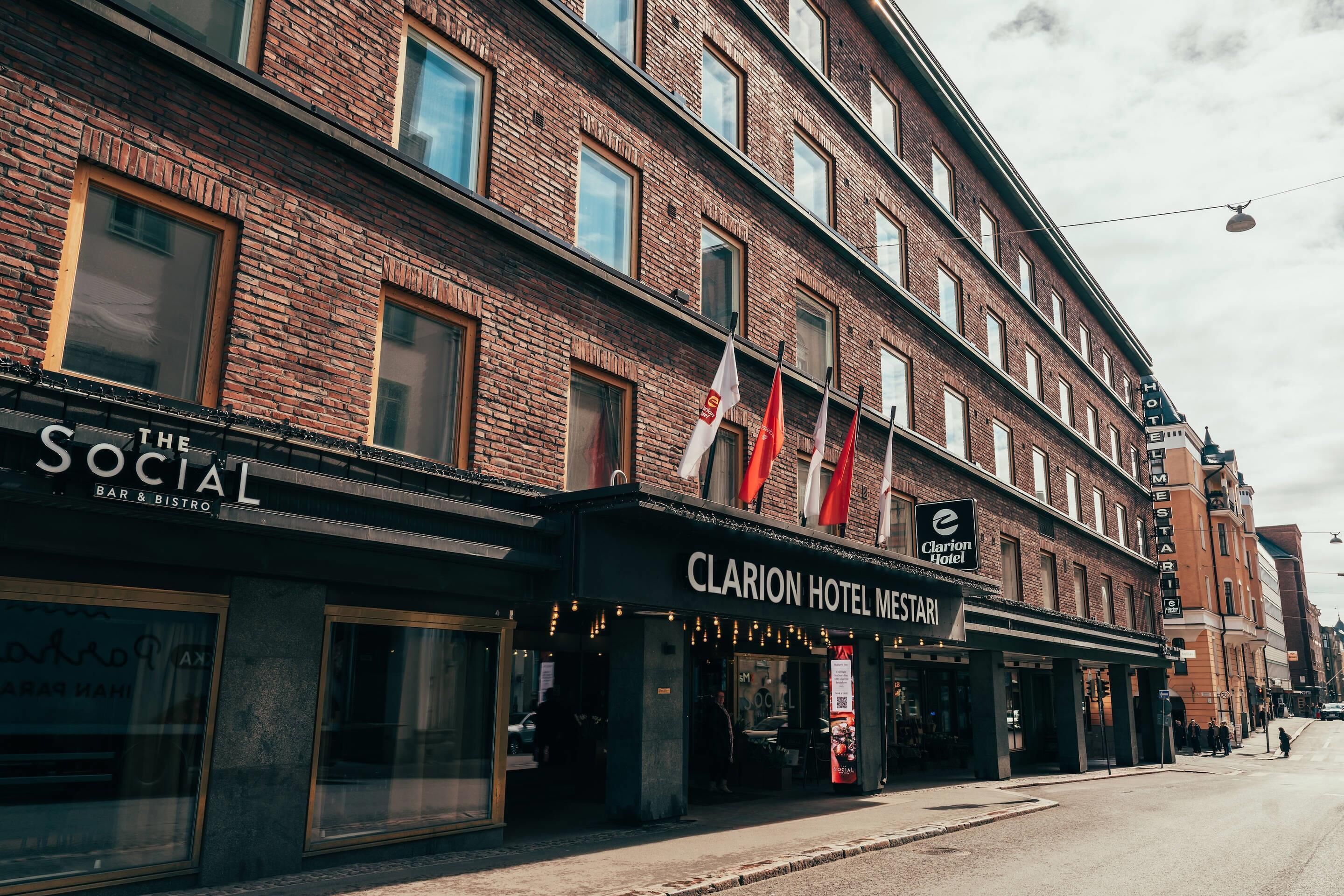 A multi-story building with a brick facade, featuring a bar and bistro named 'The Social' and a hotel named 'Clarion Hotel Mestarli' with flags above.