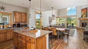 A spacious kitchen with wooden cabinets, granite countertops, and a large island. There is a dining area with a table and chairs, and a fireplace with a stone surround. Large windows offer views of the outdoors. - Mountain Willow At Grand Park 4 Bedroom Home by RedAwning (Fraser)