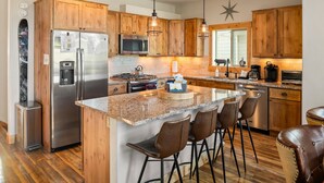 A modern kitchen with wooden cabinets, stainless steel appliances, a granite countertop, and bar stools. - Mountain Willow At Grand Park 4 Bedroom Home by RedAwning (Fraser)