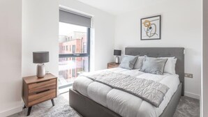 Ein modernes Schlafzimmer mit einem großen Bett, zwei Nachttischen und einem Fenster mit Blick auf Gebäude.