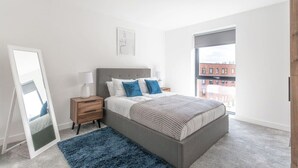 Ett modernt sovrum med en stor säng, ett nattduksbord, en spegel och utsikt över en stadssiluett genom ett stort fönster.