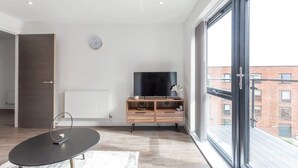 Ett modernt vardagsrum med ett stort fönster, en platt-TV placerad på ett trästativ och ett minimalistiskt soffbord.