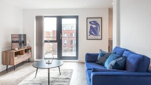 Ett modernt vardagsrum med en blå soffa, en byrå i trä, ett runt soffbord och ett stort fönster med utsikt över byggnader.