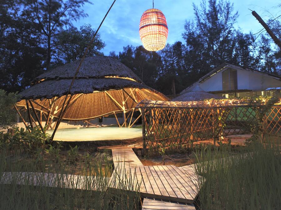 Una estructura con techo de paja y una terraza de madera que la conecta, rodeada de vegetación.