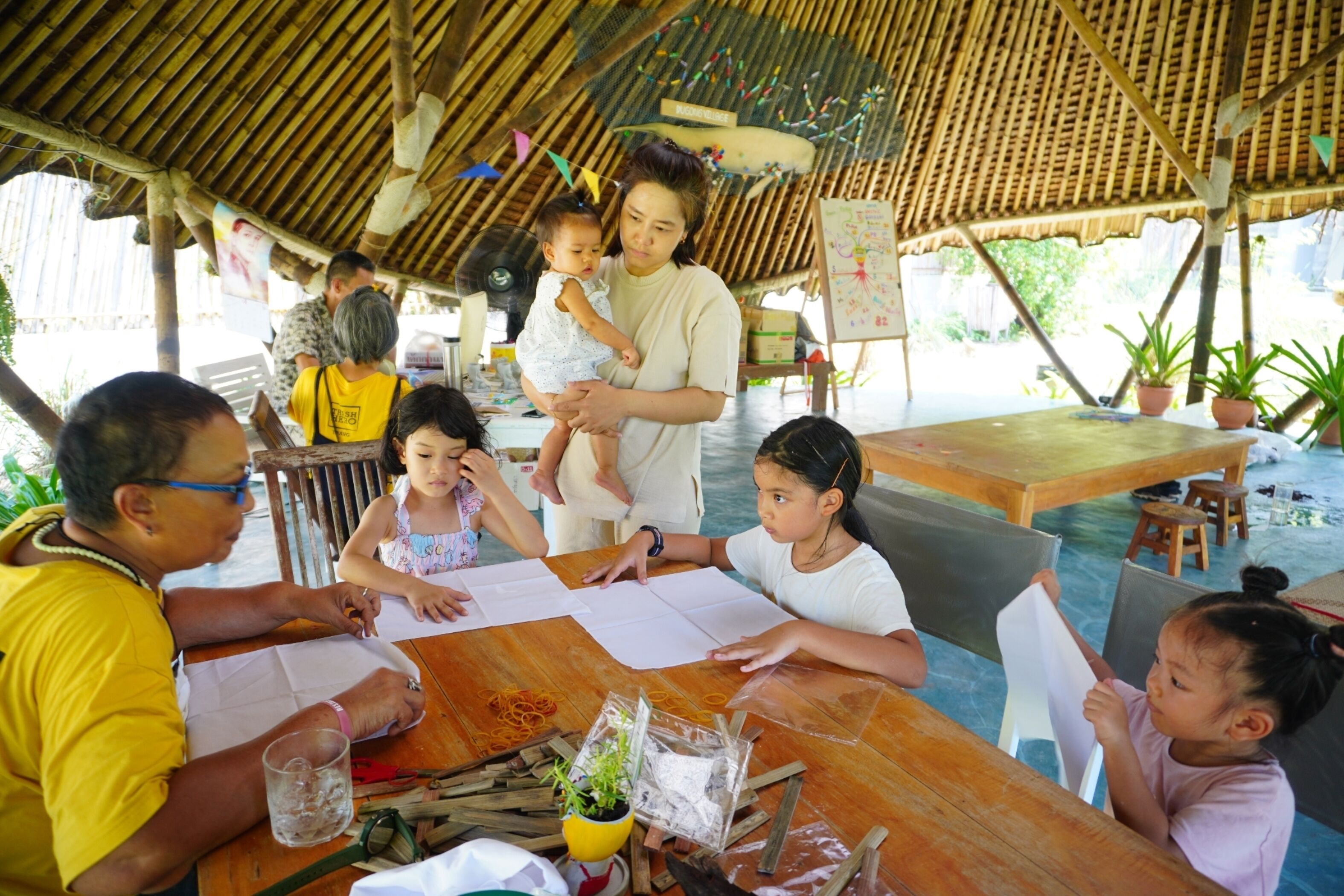 A group of people engaged in a craft activity under a thatched roof structure with tables and chairs.