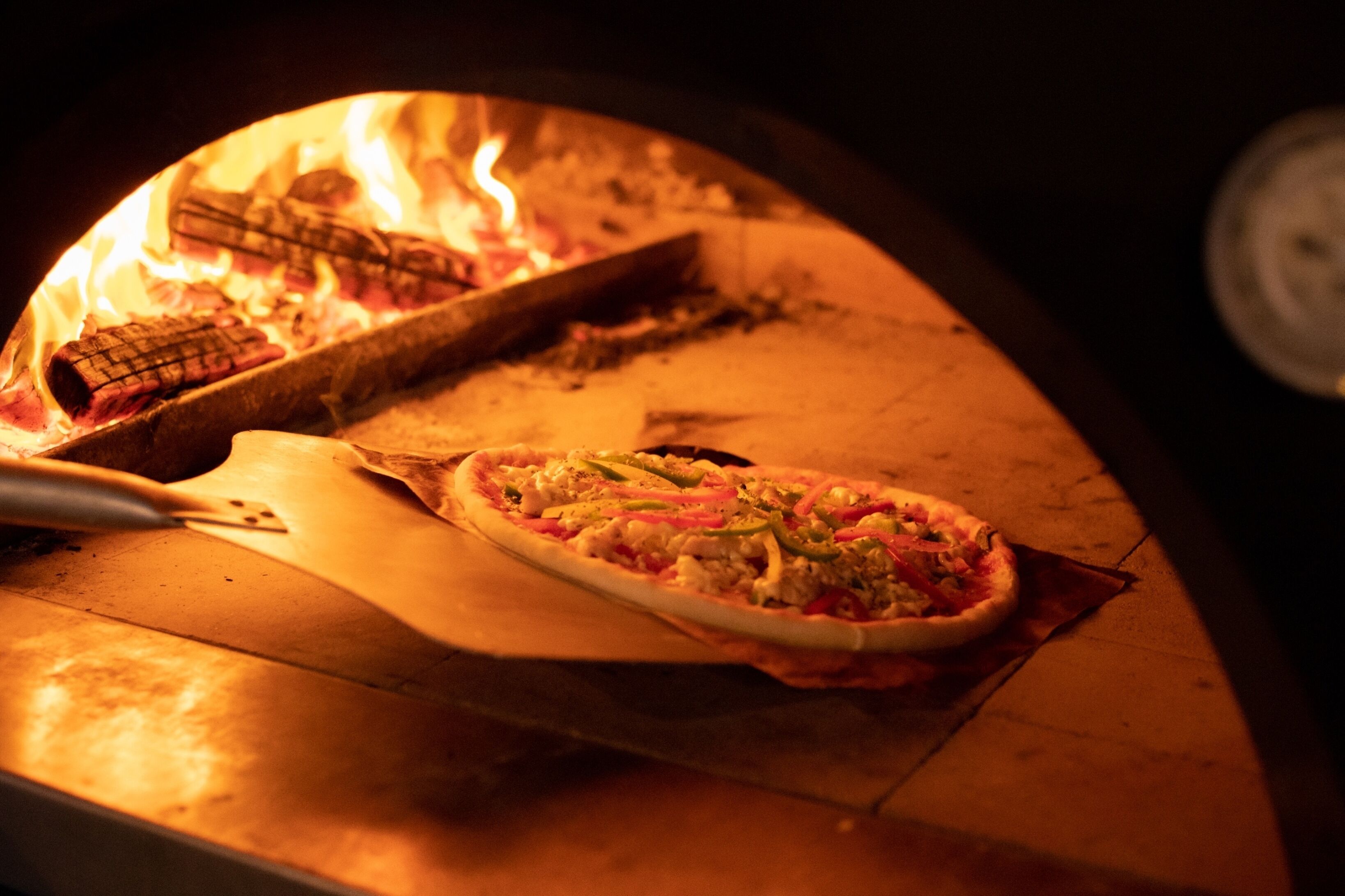 Un horno de pizza con una pizza adentro, una pala para pizza y fuego encendido.