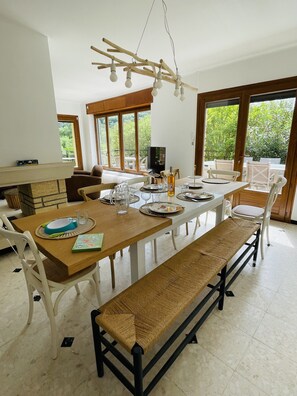 A dining area with a long wooden table, white chairs, and a bench. There is a television on a stand and a window with a view of greenery. - villa mimi (Laval-Saint-Roman)