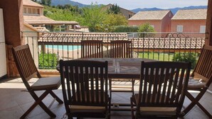 A covered patio with a table and chairs, overlooking a swimming pool and a residential area with houses and mountains in the background. - 3 bedroom apartment with swimming pool 3km from St Florent  (Oletta)