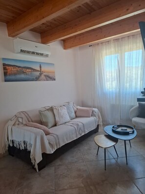Uma sala de estar com sofá, mesa redonda pequena e pintura de uma paisagem costeira.