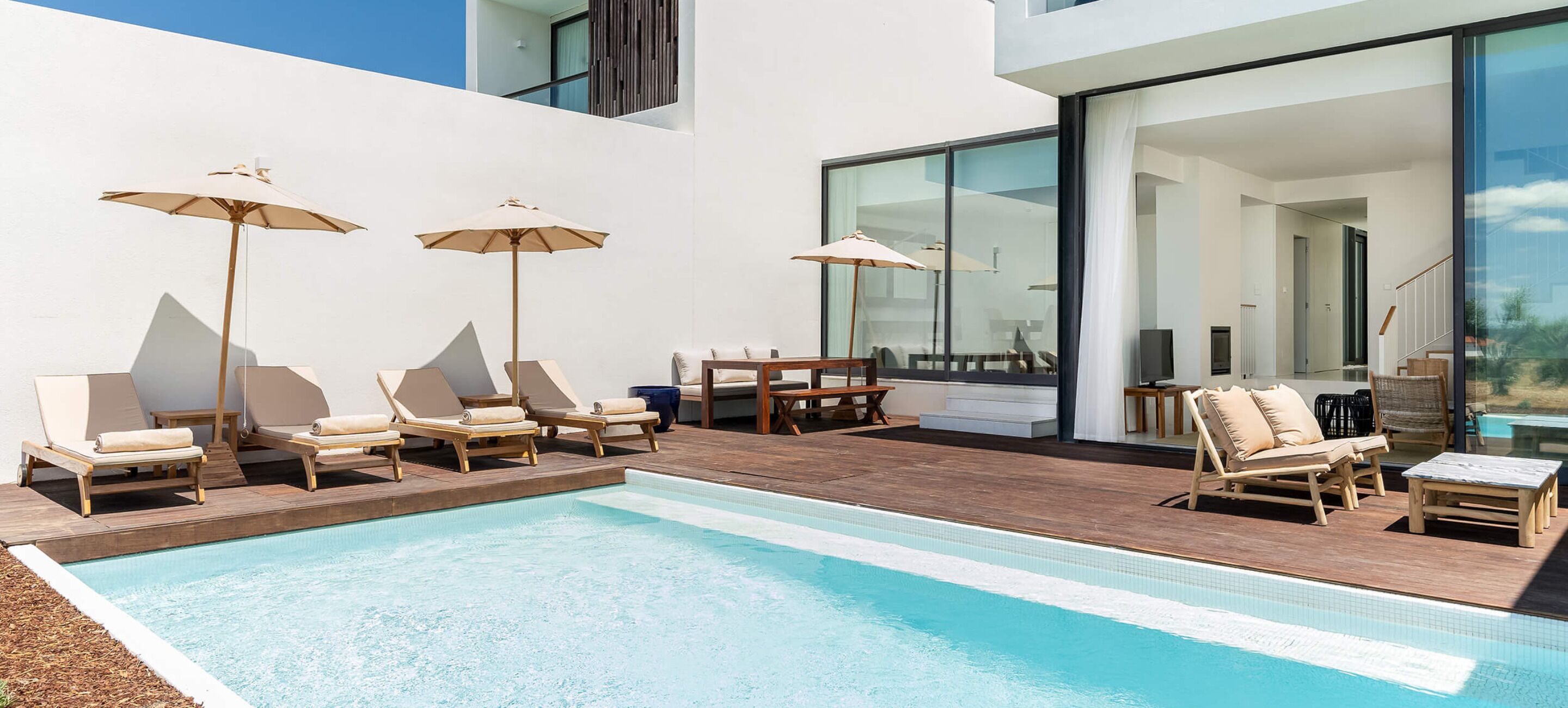Et moderne poolområde med liggestole og parasoller, en træterrasse og en glasomkranset opholdsstue.