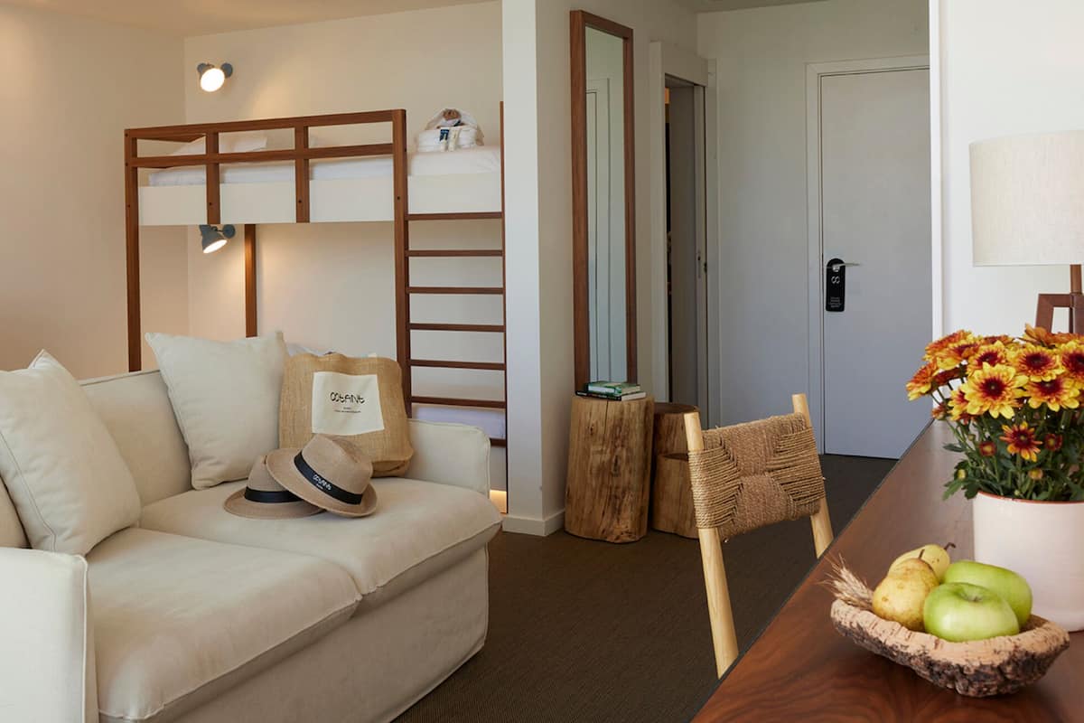 Ein Hotelzimmer mit einem Bett, einem Sofa, einem kleinen Tisch mit Äpfeln und Blumen, einer Holzleiter und einer Tür.