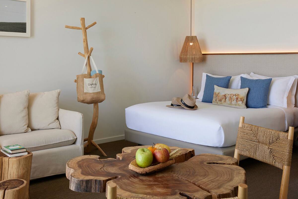 Ein gemütliches Zimmer mit einer Couch, einem Bett mit blau-weißen Kissen, einem Holz-Kleiderständer und einem Holztisch mit Früchten.