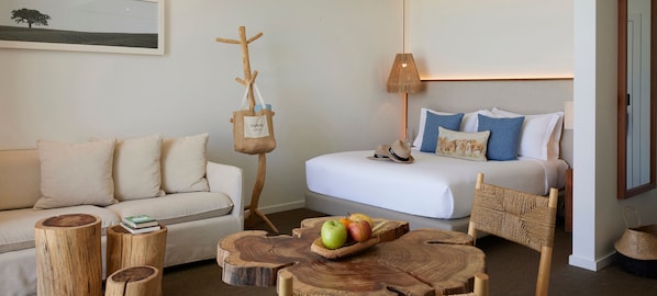 Une pièce chaleureuse avec un canapé, un lit agrémenté d’oreillers bleus et blancs, un porte-manteau en bois et une table en bois sur laquelle sont disposés des fruits.