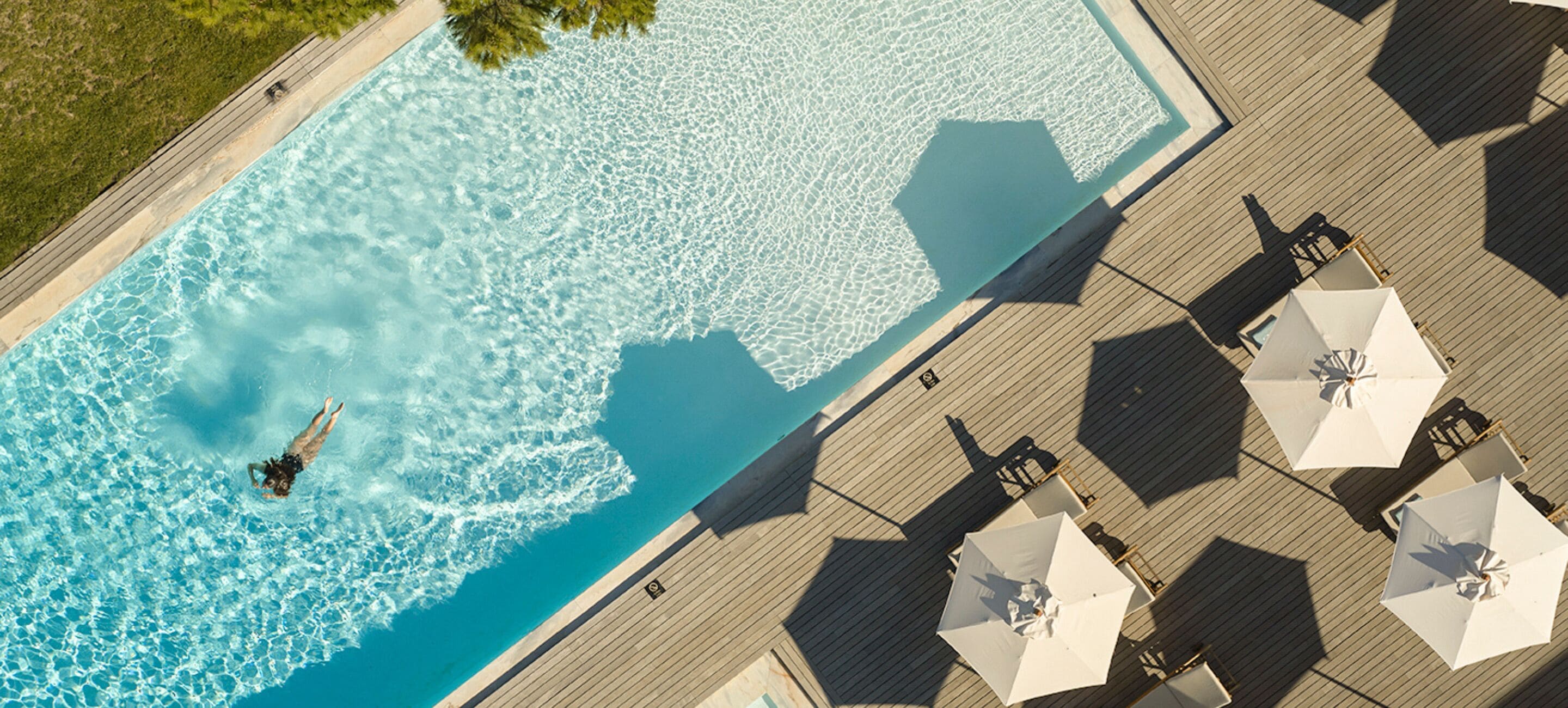An aerial view of a swimming pool with a person swimming, surrounded by sun umbrellas and deck chairs.