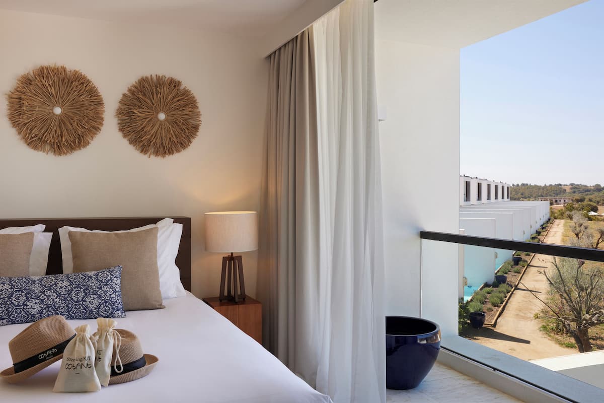 Ein Hotelzimmer mit Bett, Kissen, einer Lampe, einem Hut und Blick ins Freie.
