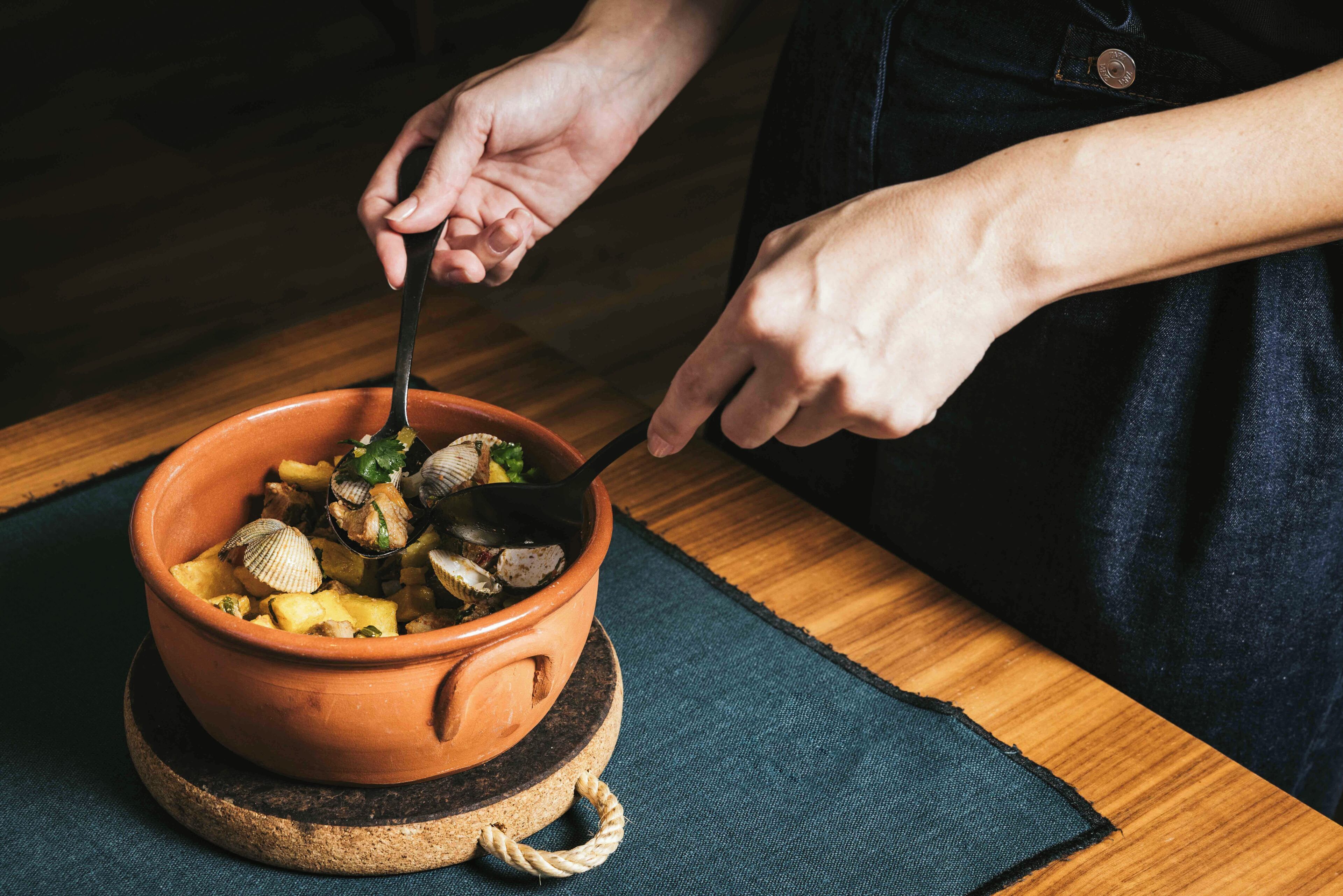 A person serving food from a terracotta bowl on a wooden table with a dark cloth underneath.