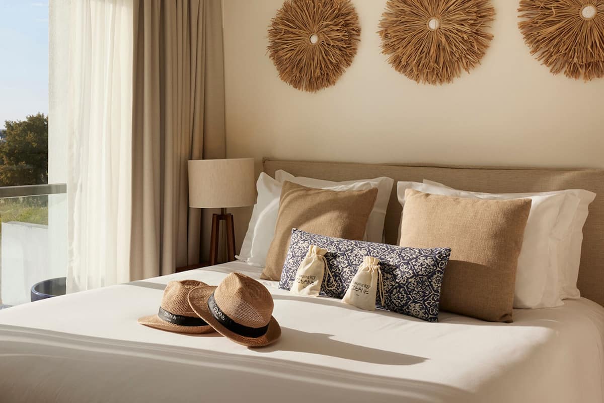 Ein Hotelzimmer mit großem Fenster, einem Bett mit vielen Kissen und einem Strohhut auf dem Bett.