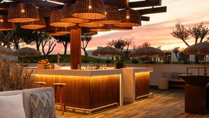 Zona de bar con mostradores de madera y taburetes, rodeada de sombrillas de paja y luces colgantes de mimbre.