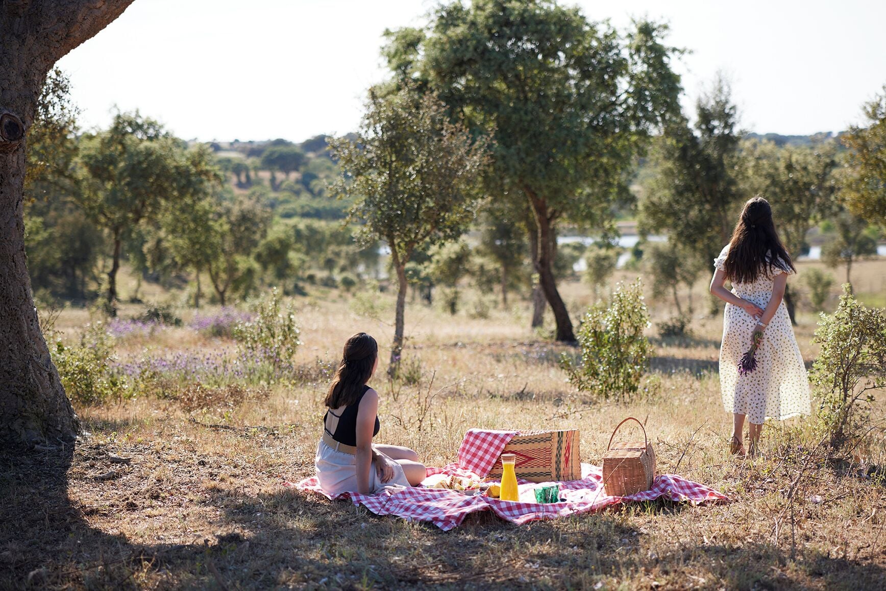 Twee personen zitten en staan op een picknickdeken in een natuurlijke omgeving, met bomen en een heldere lucht.