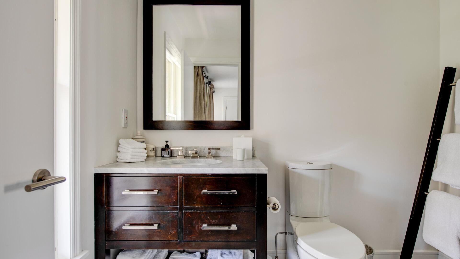 Een badkamer met een wit toilet, een donker houten wastafelmeubel met lades, een witte wasbak en een spiegel boven het meubel.