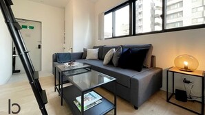 A modern living room with a sofa, a glass coffee table, a side table with a lamp, and a window with a view of buildings.