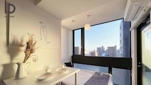 A modern hotel room with a bed, a small desk with a chair, a window with a city view, and a wall-mounted air conditioner. - b hotel Neko Yard | Modern City-Center Studio (Naka Ward, Hiroshima)