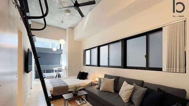 A modern hotel room with a sofa, a small table, a ladder, and a large window with blinds.
