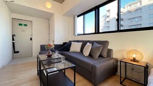 Una habitación de hotel moderna con un sofá, una mesa de centro de vidrio, una mesita de noche con lámpara y vistas a edificios a través de amplias ventanas.