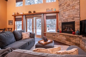A cozy living room with a stone fireplace, a gray sofa, and a large window overlooking a snowy landscape. - Bigfork Family Home | Hot tub & Playset (Bigfork)