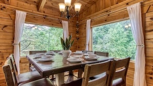 A wooden cabin interior with a dining table set for four, surrounded by chairs, and large windows offering a view of the forest. - Dreamy Forest Home, 2 Decks, Hot Tub, Outdoor Pool | 10 Mins to Broadway, Perfect for Groups, 24 Hour Hospitality, Sleep 10 w/ Wraparound Deck, Scenic Views & Access to Resort Pool,  Near The Parkway, Tanger Outlets & Dollywood (Pigeon Forge)