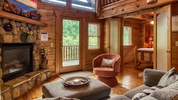 A log cabin interior with a stone fireplace, a cozy seating area, and a balcony with a wooden railing. - Dreamy Forest Home, 2 Decks, Hot Tub, Outdoor Pool | 10 Mins to Broadway, Perfect for Groups, 24 Hour Hospitality, Sleep 10 w/ Wraparound Deck, Scenic Views & Access to Resort Pool,  Near The Parkway, Tanger Outlets & Dollywood (Pigeon Forge)