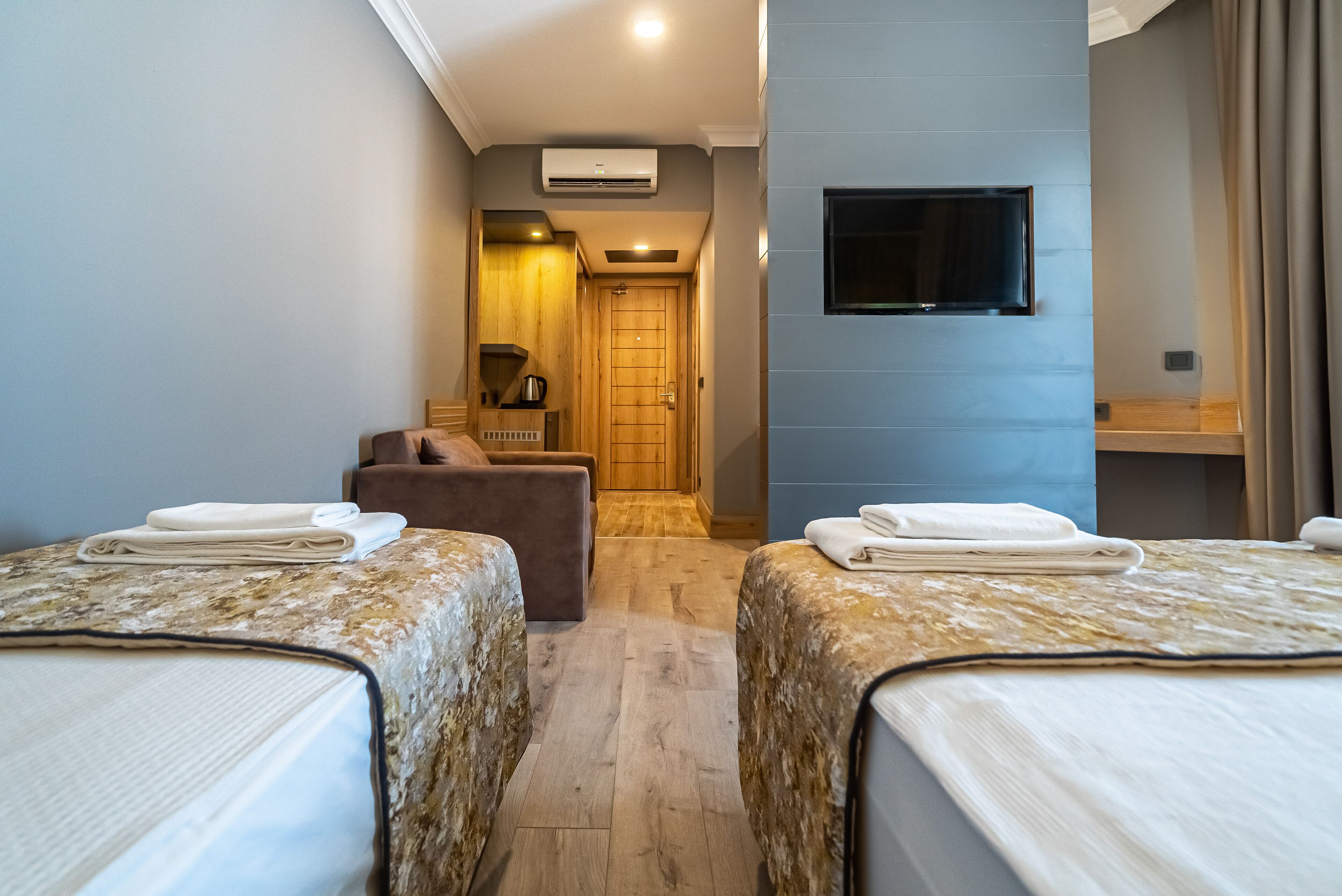 A hotel room with two beds, a wooden floor, a TV mounted on the wall, and a small seating area with a chair and a table.