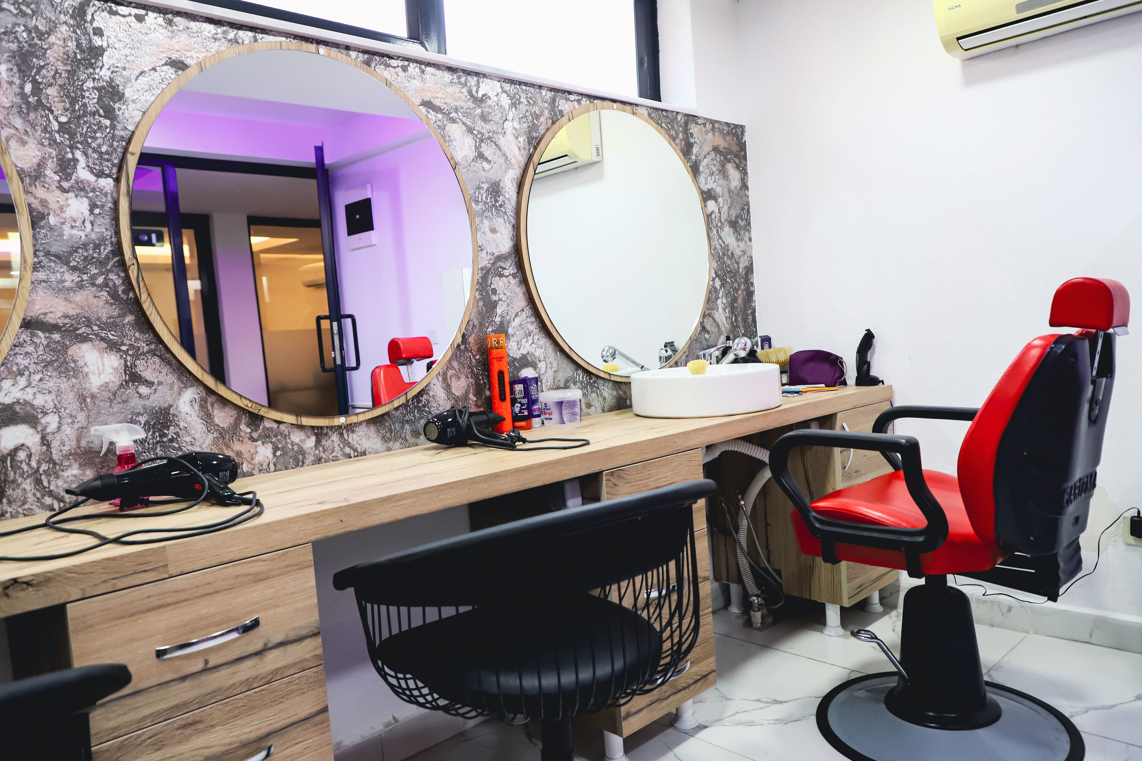A salon with a wooden counter, two large mirrors, a red and black salon chair, and a black chair with a mesh backrest.