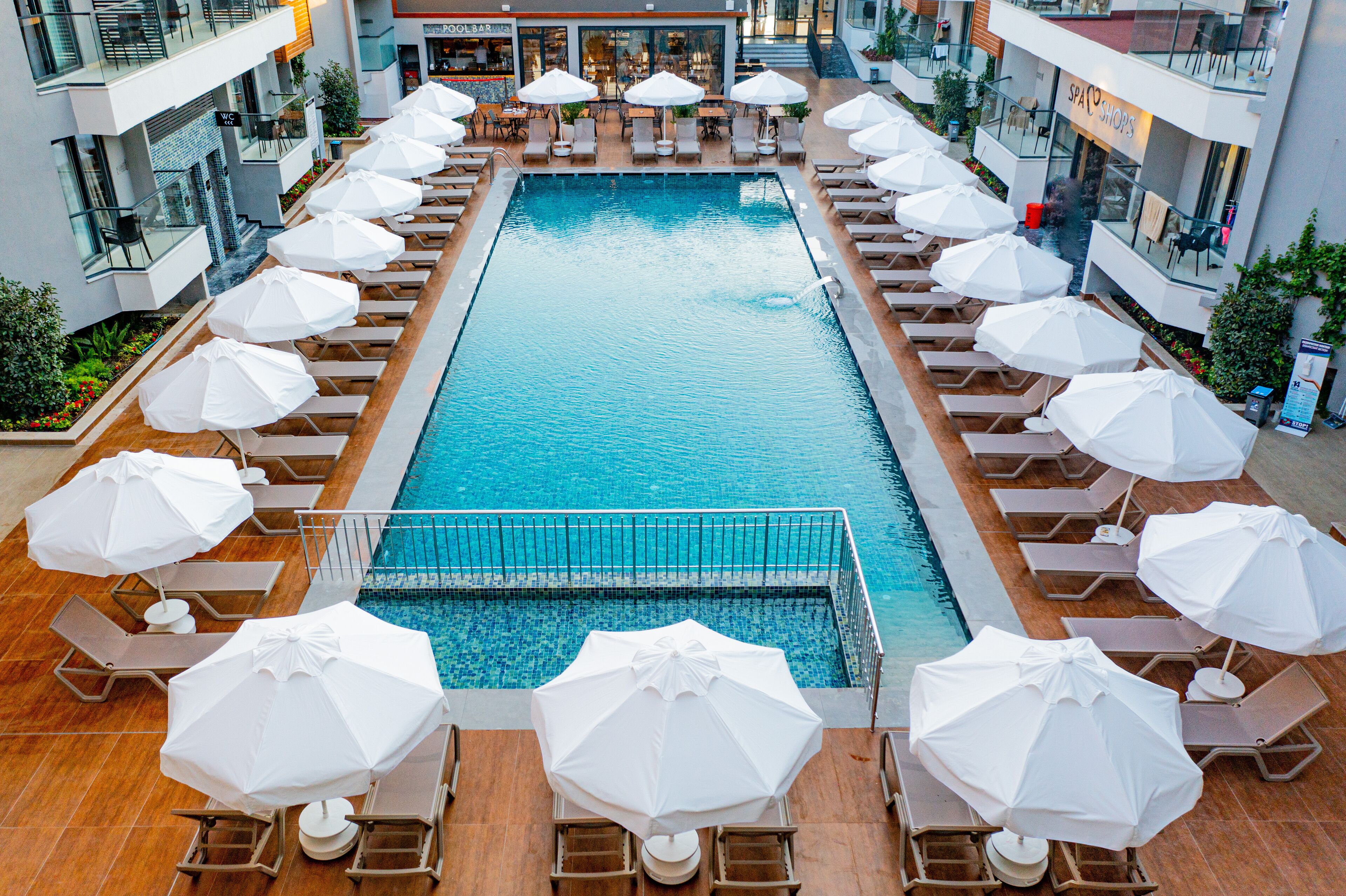 A pool area with sun loungers and umbrellas, surrounded by buildings with shops and a spa.