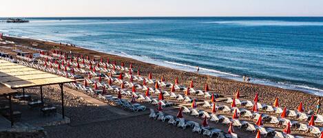 A beach with rows of sun loungers and umbrellas, a pier in the background, and a clear sky.