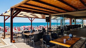 Un restaurante frente a la playa con terraza, vistas al mar y una zona cubierta con mesas y sillas.