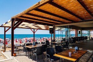 A beachside restaurant with outdoor seating, a view of the sea, and a covered area with tables and chairs.