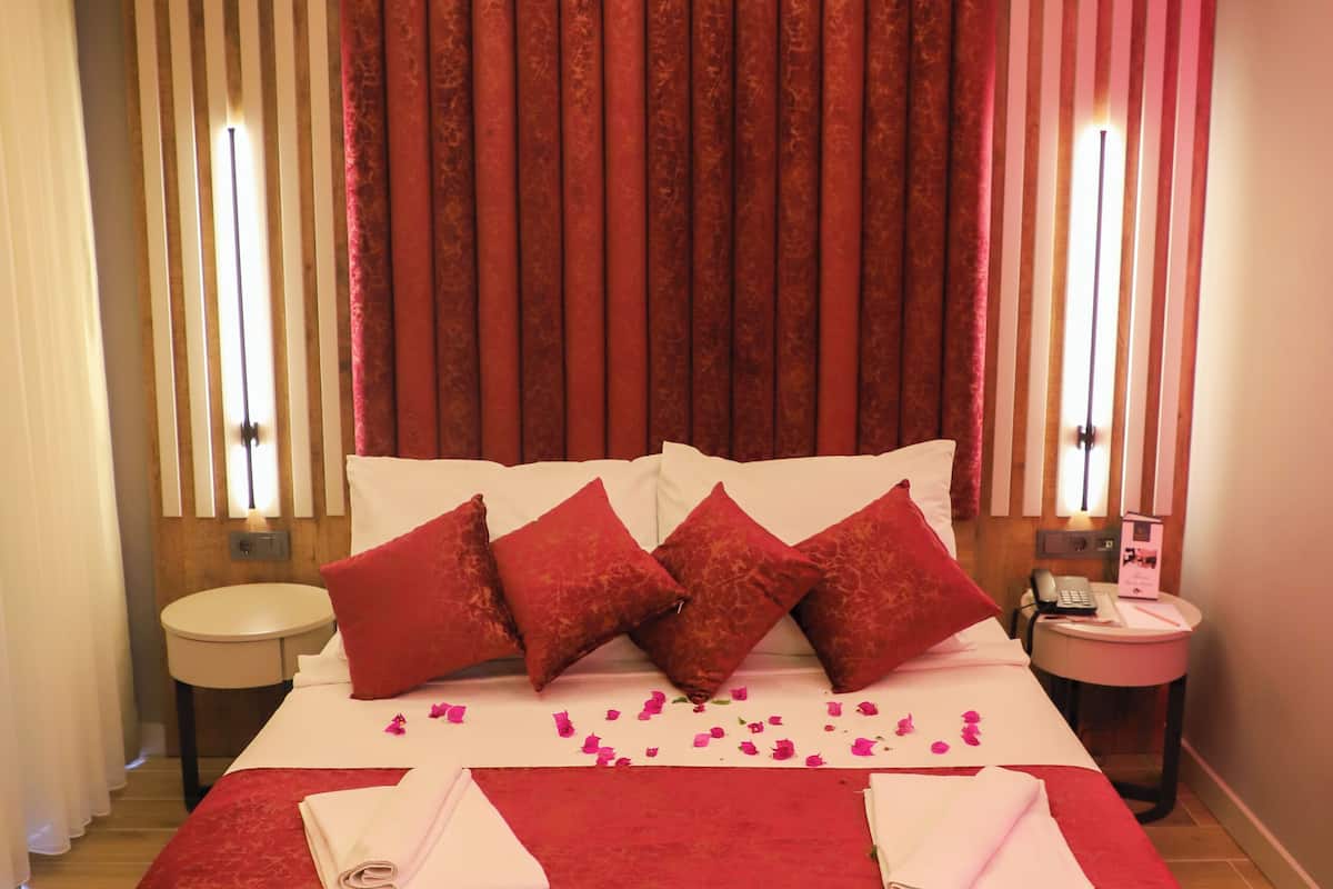 A hotel room with a large bed, red curtains, a bedside table with a lamp, and a small round table with a phone and a menu.