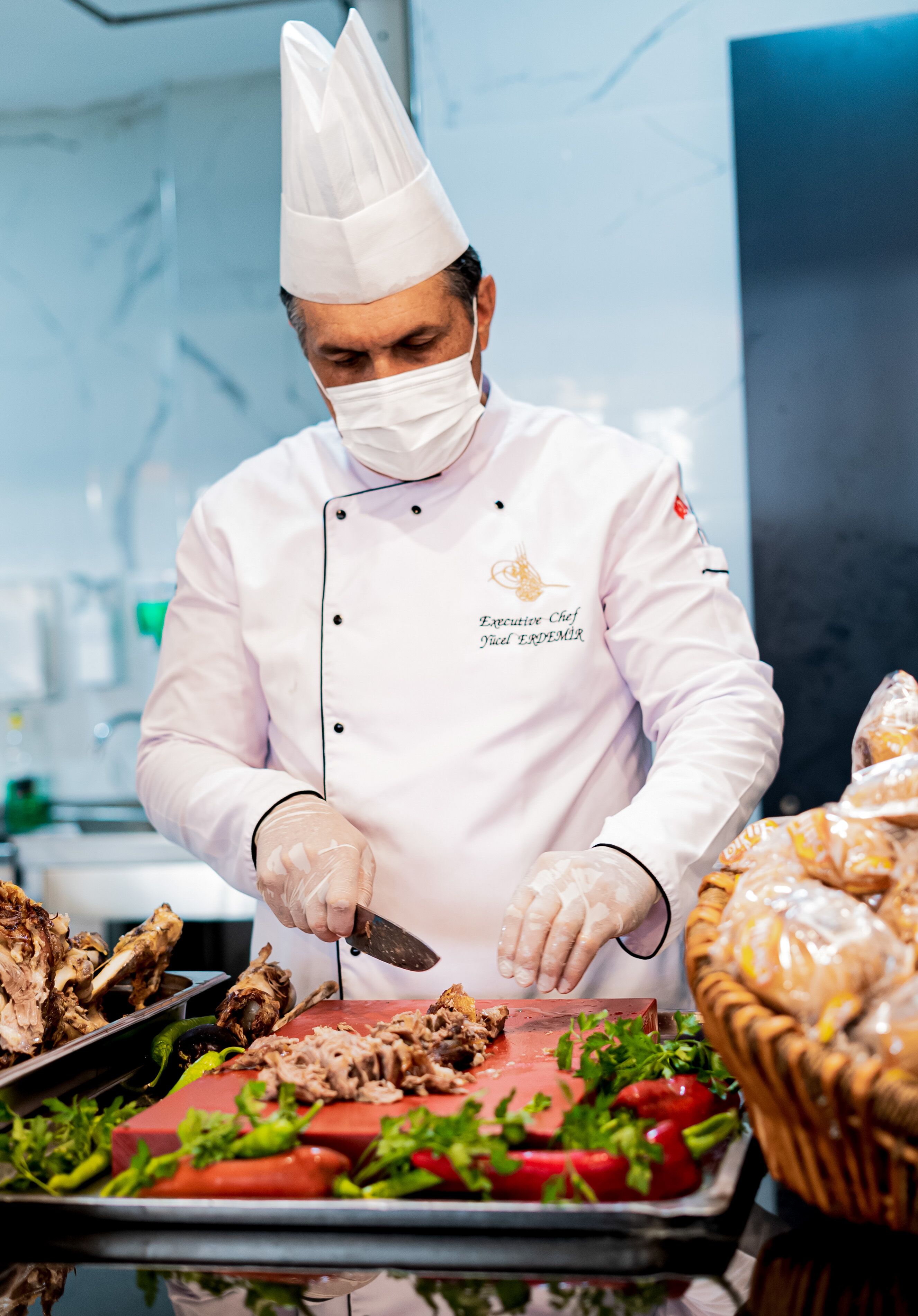 Un chef con uniforme blanco y gorro de cocinero cortando carne en una bandeja con verduras.