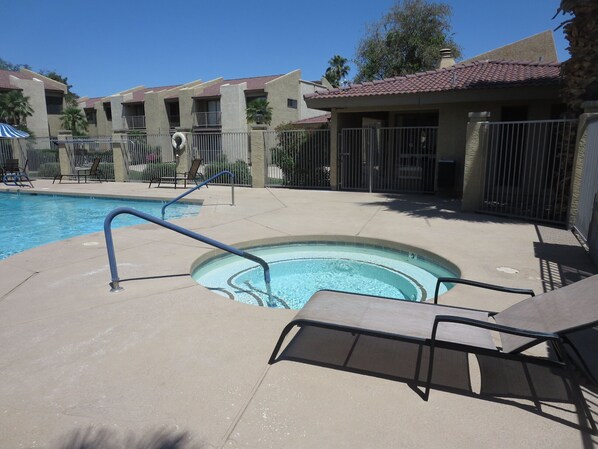 A swimming pool with a hot tub, lounge chairs, and a fenced area with a building in the background.