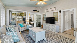 A cozy living room with a sofa, armchair, and a TV on a cabinet. There is a ceiling fan and a sign that says "Come in and stay awhile." - 3 bedroom, short walk to the beach with an amazing game room! (Hilton Head Island)