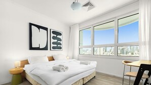 Una habitación de hotel moderna con una cama grande, una mesita de madera, una silla y vistas a la ciudad a través de amplios ventanales.