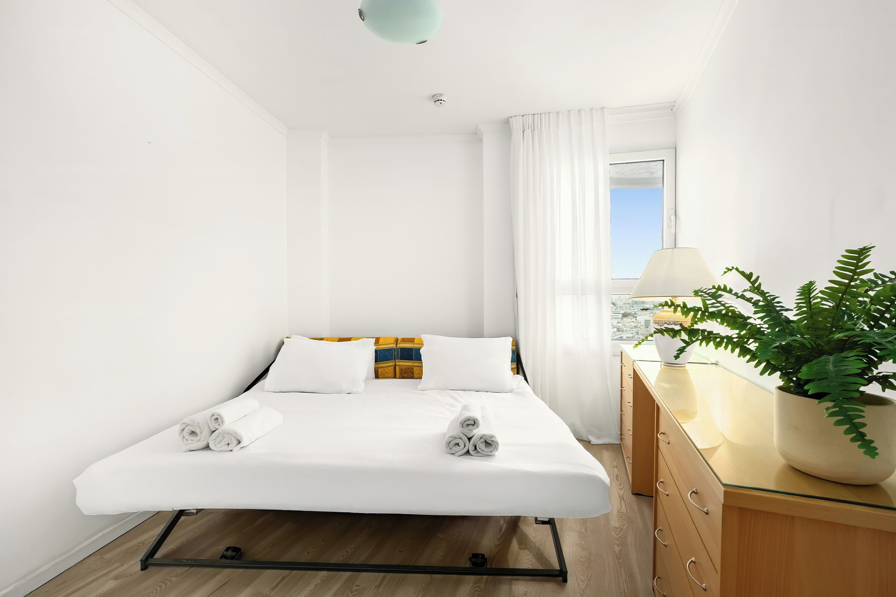 A modern hotel room with a bed, a wooden dresser, a potted plant, and a view of the city through the window.