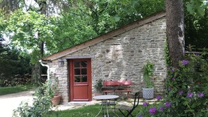 A stone building with a red door, a small patio with a table and chairs, and surrounding greenery.