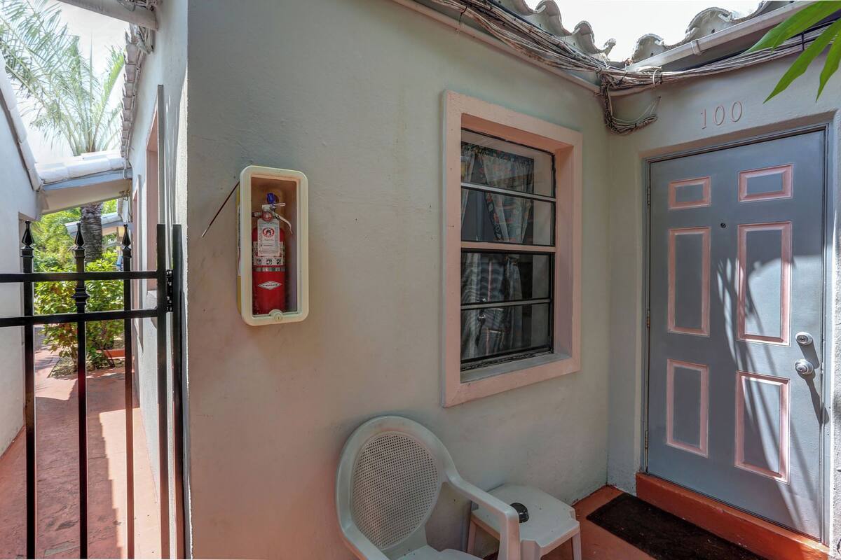 A building entrance with a fire extinguisher mounted on the wall, a window with curtains, a door with a number plate, and a chair outside.