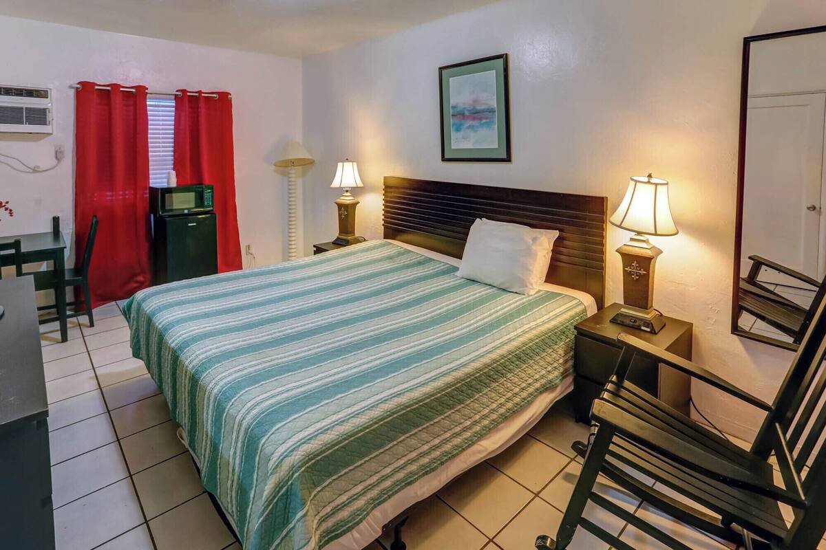 A hotel room with a bed, bedside lamps, a desk with a computer, a chair, and a red curtain.