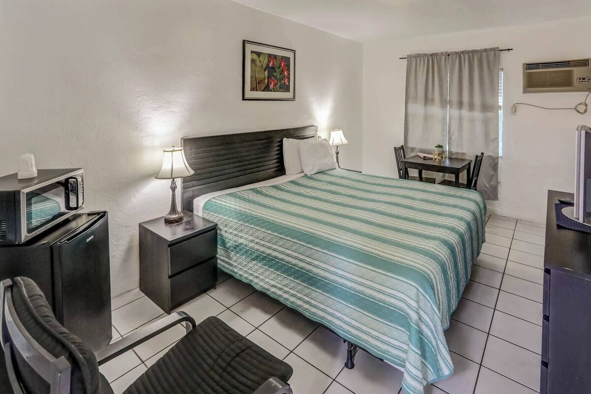 A hotel room with a bed, a desk, a chair, a table, a TV, and a lamp.