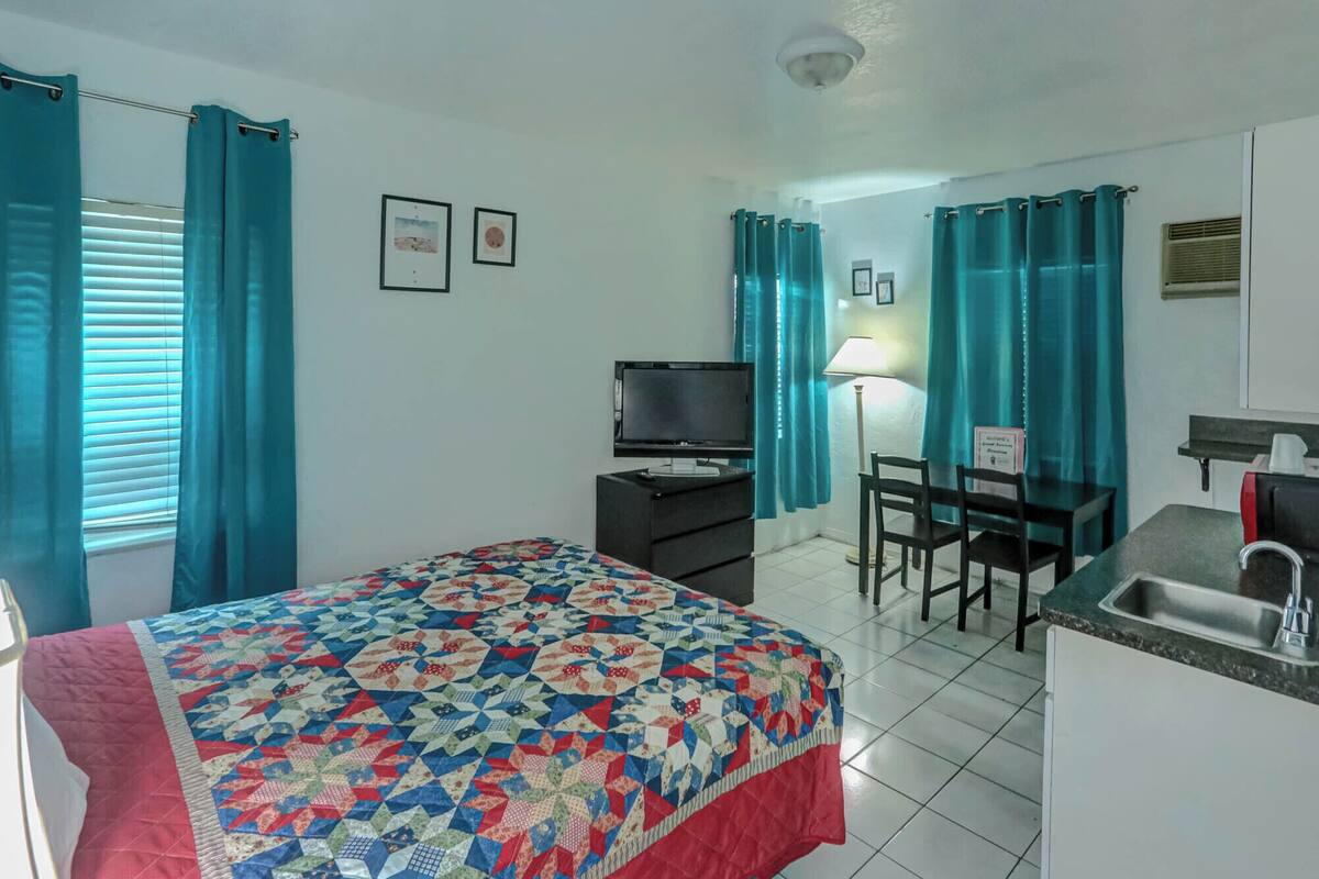 A hotel room with a bed, a desk with a chair, a television, a sink, and a window with curtains.